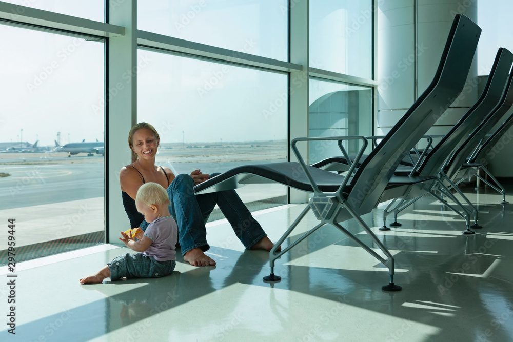 Mother with little kid wait for boarding to plane flight in airport transit hall. Baby look through the window at flying airplane. Active family lifestyle, travel by air with child on summer vacation.