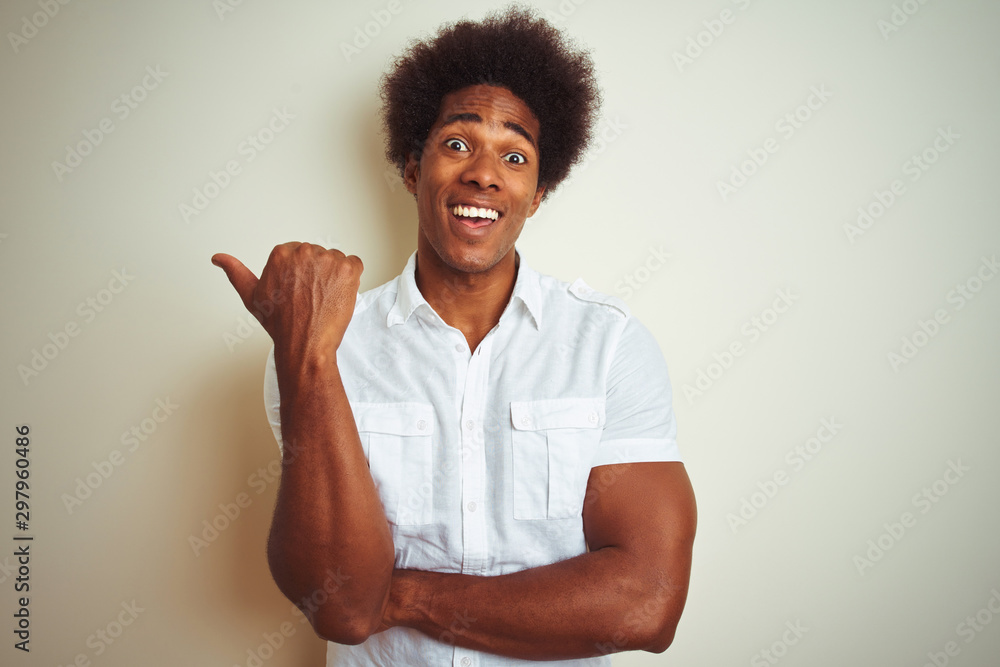 African american man with afro hair wearing shirt standing over isolated white background smiling with happy face looking and pointing to the side with thumb up. African american man with afro hair wearing shirt standing over isolated white background smiling with happy face looking and pointing to the side with thumb up.