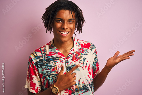 Afro man with dreadlocks on vacation wearing floral shirt over isolated pink background amazed and smiling to the camera while presenting with hand and pointing with finger.