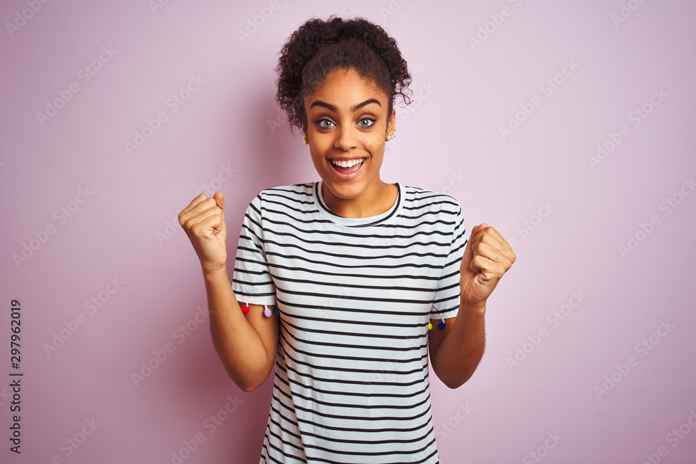 African american woman wearing navy striped t-shirt standing over isolated pink background celebrating surprised and amazed for success with arms raised and open eyes. Winner concept. African american woman wearing navy striped t-shirt standing over isolated pink background celebrating surprised and amazed for success with arms raised and open eyes. Winner concept.
