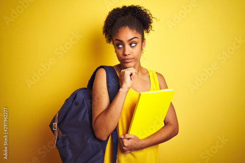 American student woman wearing backpack holding notebook over isolated yellow background serious face thinking about question, very confused idea