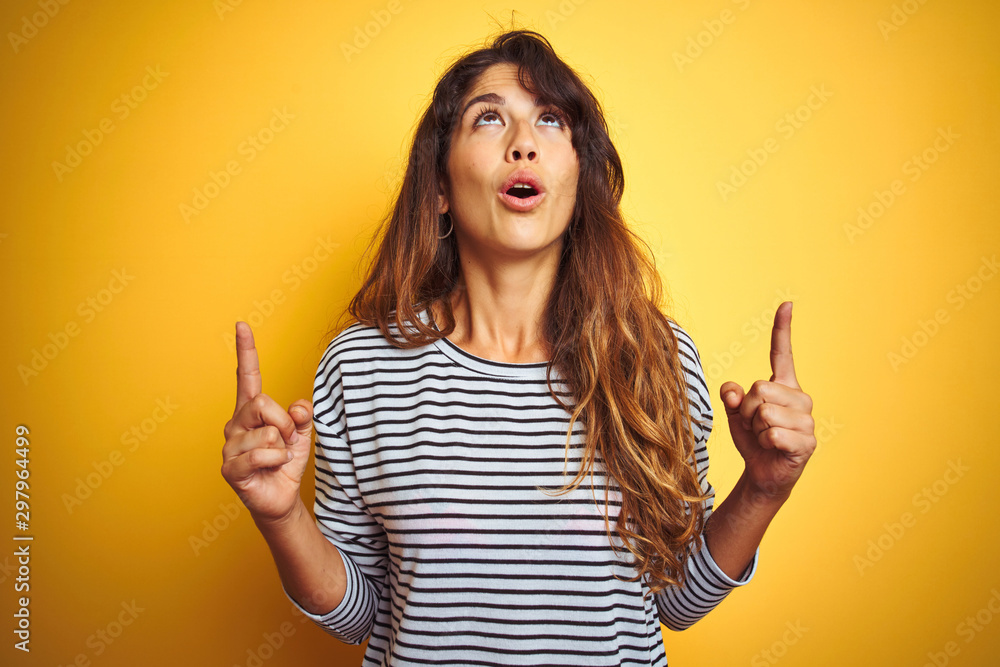 Young beautiful woman wearing stripes t-shirt standing over yelllow isolated background amazed and surprised looking up and pointing with fingers and raised arms. Young beautiful woman wearing stripes t-shirt standing over yelllow isolated background amazed and surprised looking up and pointing with fingers and raised arms.