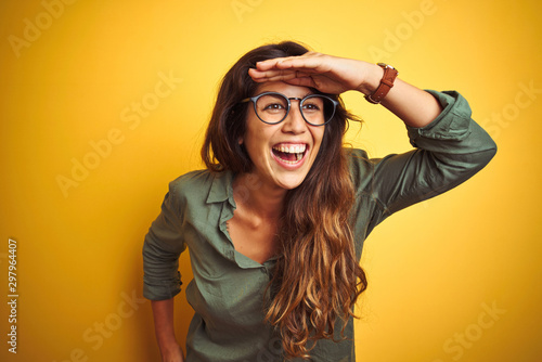 Young beautiful woman wearing green shirt and glasses over yelllow isolated background very happy and smiling looking far away with hand over head. Searching concept. photo