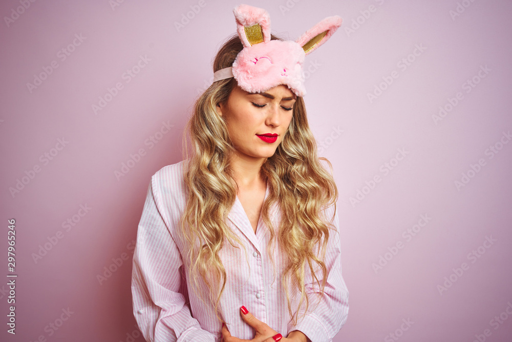 Young beautiful woman wearing pajama and sleep mask over pink isolated background with hand on stomach because nausea, painful disease feeling unwell. Ache concept.