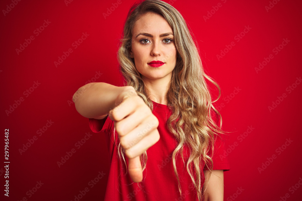 Young beautiful woman wearing basic t-shirt standing over red isolated background looking unhappy and angry showing rejection and negative with thumbs down gesture. Bad expression. Young beautiful woman wearing basic t-shirt standing over red isolated background looking unhappy and angry showing rejection and negative with thumbs down gesture. Bad expression.