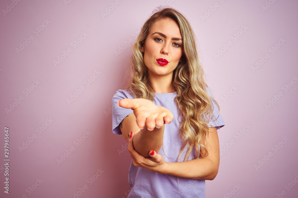 Young beautiful woman wearing purple t-shirt standing over pink isolated background looking at the camera blowing a kiss with hand on air being lovely and sexy. Love expression. Young beautiful woman wearing purple t-shirt standing over pink isolated background looking at the camera blowing a kiss with hand on air being lovely and sexy. Love expression.