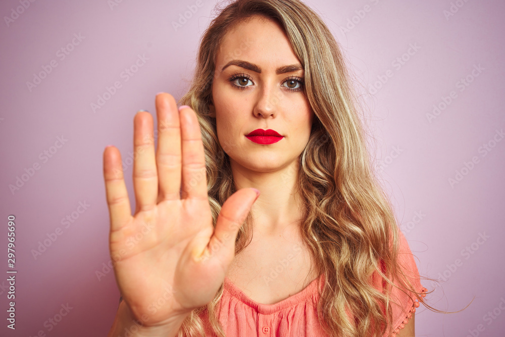 Young beautiful woman wearing t-shirt standing over pink isolated background with open hand doing stop sign with serious and confident expression, defense gesture Young beautiful woman wearing t-shirt standing over pink isolated background with open hand doing stop sign with serious and confident expression, defense gesture