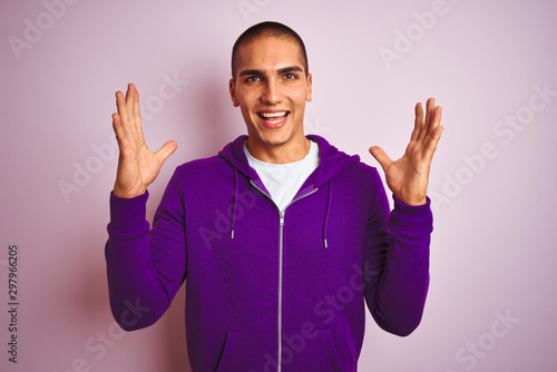 Young handsome man wearing purple sweatshirt over pink isolated background celebrating mad and crazy for success with arms raised and closed eyes screaming excited. Winner concept