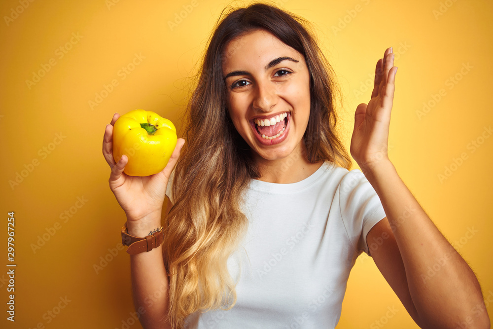 Young beautiful woman holding pepper over yellow isolated background very happy and excited, winner expression celebrating victory screaming with big smile and raised hands Young beautiful woman holding pepper over yellow isolated background very happy and excited, winner expression celebrating victory screaming with big smile and raised hands