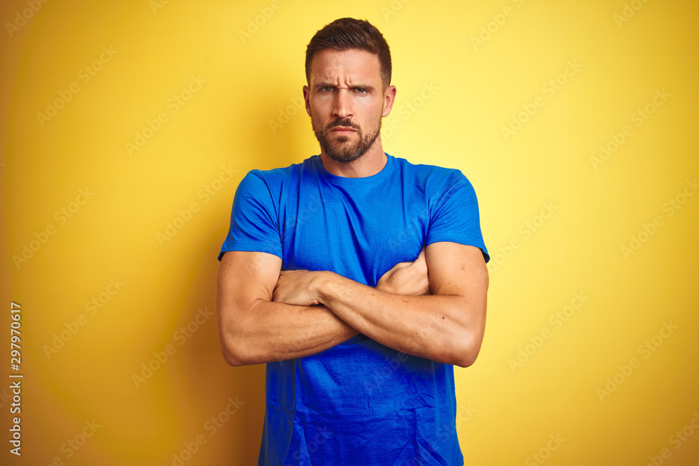 Young handsome man wearing casual blue t-shirt over yellow isolated background skeptic and nervous, disapproving expression on face with crossed arms. Negative person. Young handsome man wearing casual blue t-shirt over yellow isolated background skeptic and nervous, disapproving expression on face with crossed arms. Negative person.