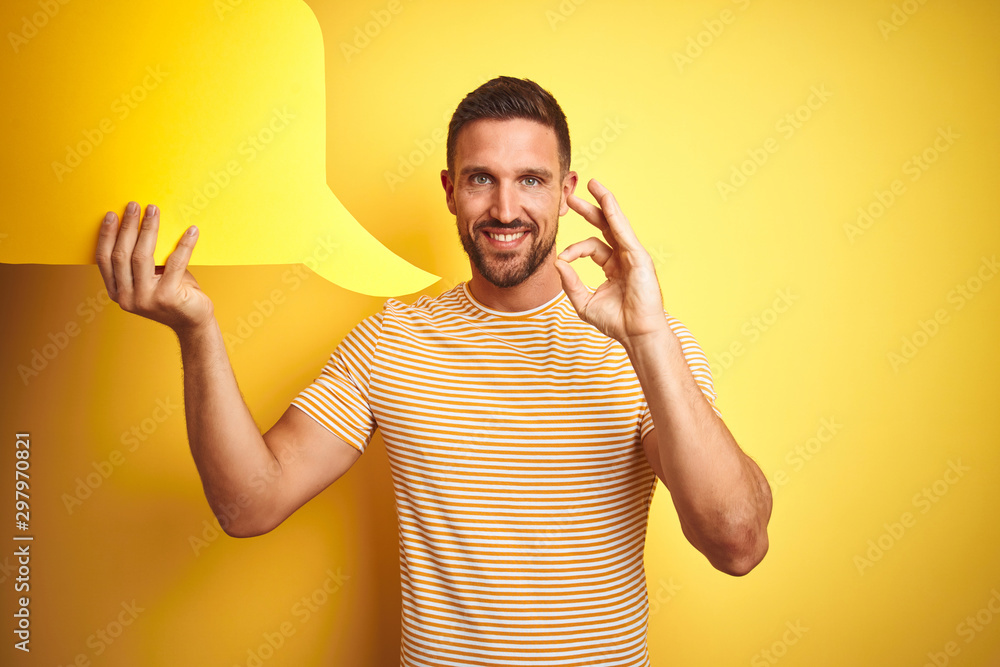 Young man holding empty speech bubble for message over yellow isolated background doing ok sign with fingers, excellent symbol Young man holding empty speech bubble for message over yellow isolated background doing ok sign with fingers, excellent symbol