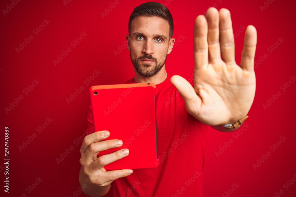 Young man using digital touchpad tablet over red isolated background with open hand doing stop sign with serious and confident expression, defense gesture Young man using digital touchpad tablet over red isolated background with open hand doing stop sign with serious and confident expression, defense gesture