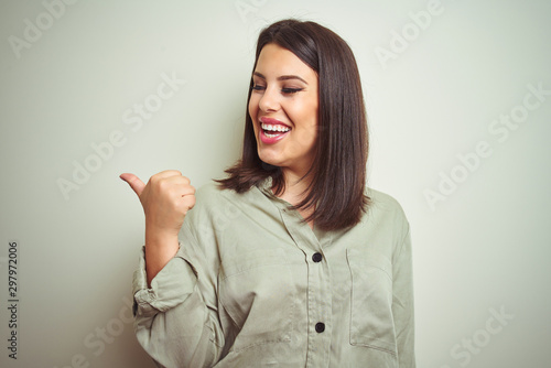Young beautiful brunette woman wearing green shirt over isolated background smiling with happy face looking and pointing to the side with thumb up.
