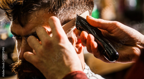 Haircut concept. Man visiting hairstylist in barbershop. Barber works with hair clipper. Hipster client getting haircut. Hands of barber with hair clipper, close up photo