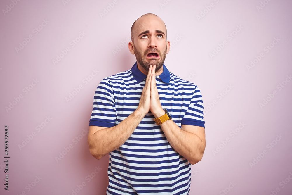 Young bald man with beard wearing casual striped blue t-shirt over pink isolated background begging and praying with hands together with hope expression on face very emotional and worried. Young bald man with beard wearing casual striped blue t-shirt over pink isolated background begging and praying with hands together with hope expression on face very emotional and worried.