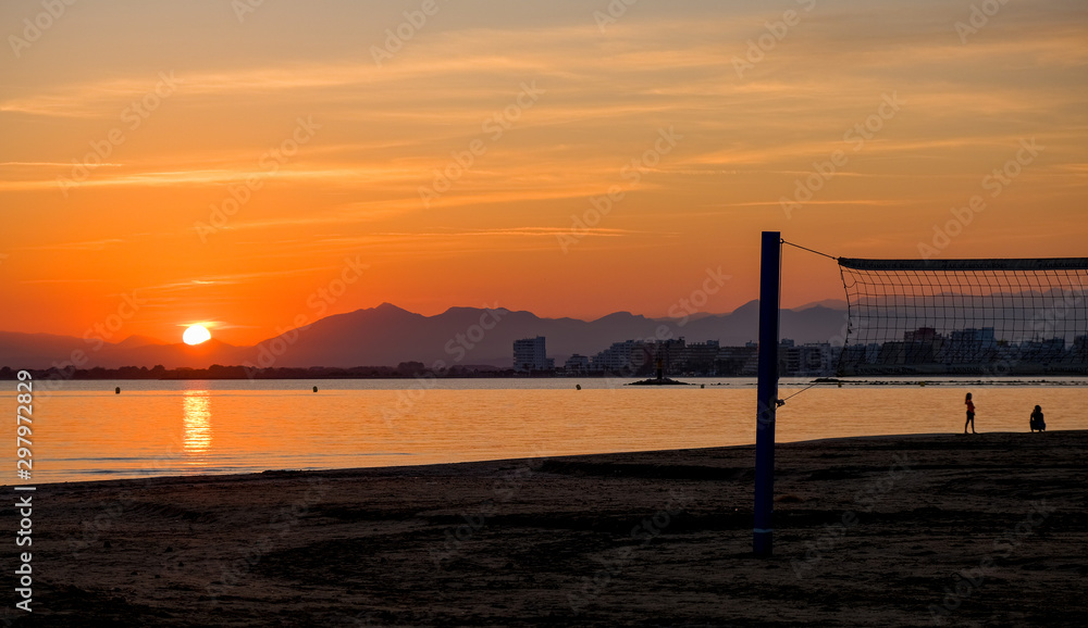 Beach volleyball court on a sandy beach by sea in evening. Amazing beautiful summer golden sunset over a bay of Roses, Catalunya, Spain. Sandy beach, calm sea on mountains and evening sky background.