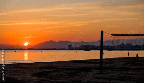Beach volleyball court on a sandy beach by sea in evening. Amazing beautiful summer golden sunset over a bay of Roses  Catalunya  Spain. Sandy beach  calm sea on mountains and evening sky background.