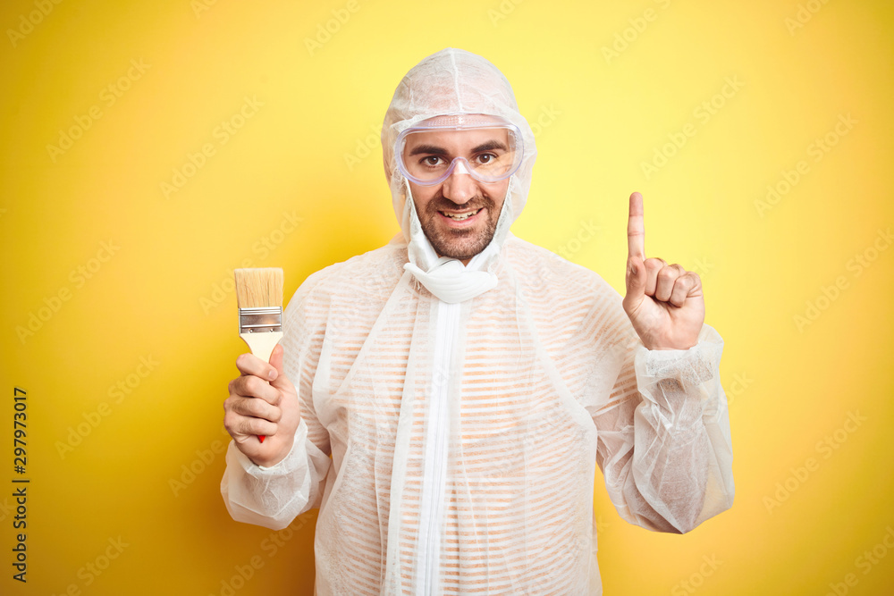 Young man wearing painter equipment and holding painting brush over isolated yellow background surprised with an idea or question pointing finger with happy face, number one Young man wearing painter equipment and holding painting brush over isolated yellow background surprised with an idea or question pointing finger with happy face, number one
