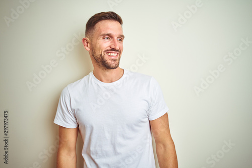 Young handsome man wearing casual white t-shirt over isolated background looking away to side with smile on face, natural expression. Laughing confident.