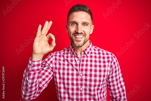 Young handsome man over red isolated background smiling positive doing ok sign with hand and fingers. Successful expression.