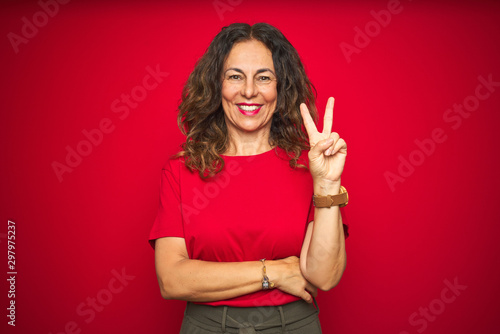 Obraz na płótnie Middle age senior woman with curly hair over red isolated background smiling with happy face winking at the camera doing victory sign