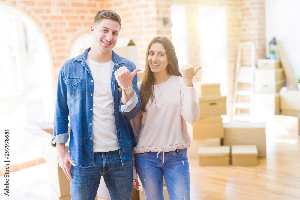 Beautiful young couple moving to a new house smiling with happy face looking and pointing to the side with thumb up. Beautiful young couple moving to a new house smiling with happy face looking and pointing to the side with thumb up.