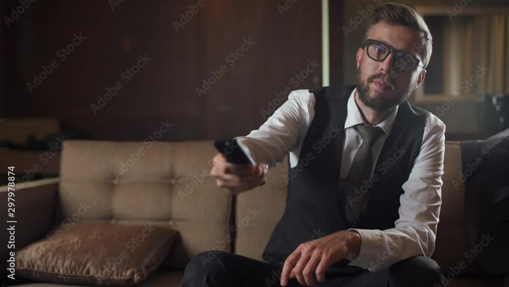 Young businessman in glasses watches the news on TV and turn on channels, man in the suit sitting on the couch in the hotel, front view, business trip. Young businessman in glasses watches the news on TV and turn on channels, man in the suit sitting on the couch in the hotel, front view, business trip.