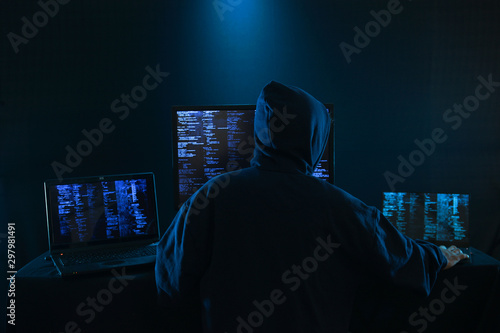 Rear View Of A Hacker Using Multiple Computers For Stealing Data On Desk