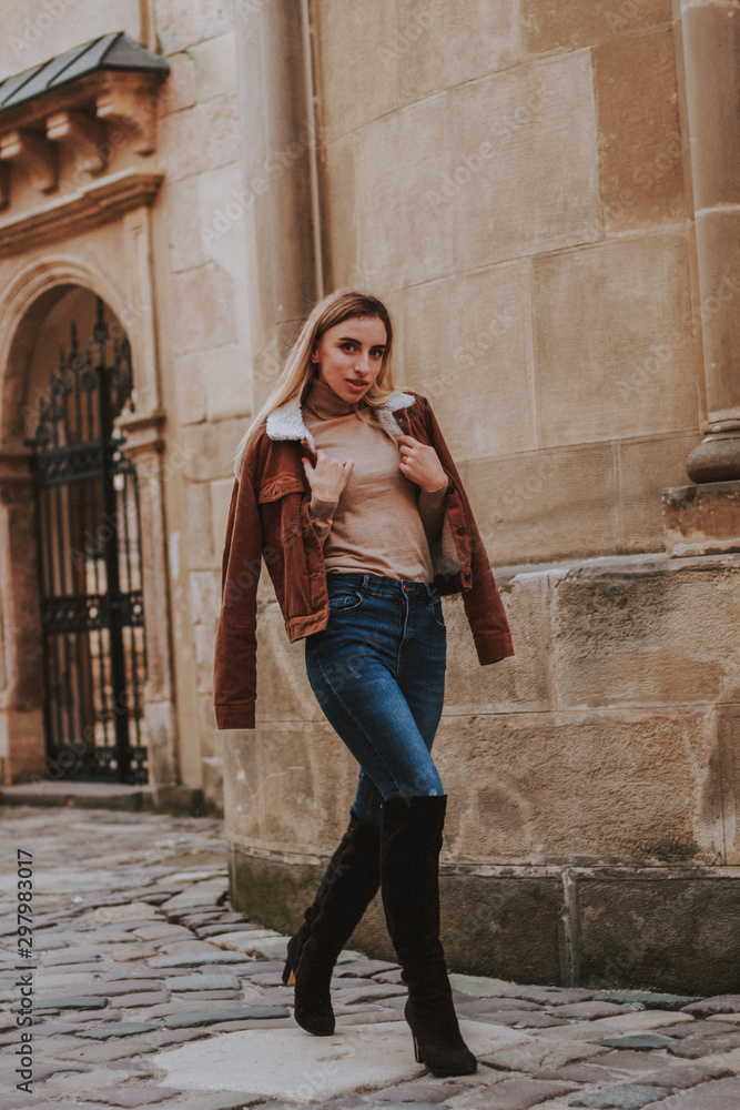 Stylish autumn fashionable image of a girl. Brown jacket with fur, jeans, boots, basic beige golf. Blonde on old city background