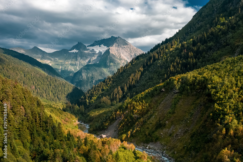 Fototapeta premium Panoramic view of Mount Tsakhvoa, autumn forest on steep slopes, curved river valley foreground; crones of pine fir trees in warm sunset light; dramatic clouds on snowy peak; natural travel background