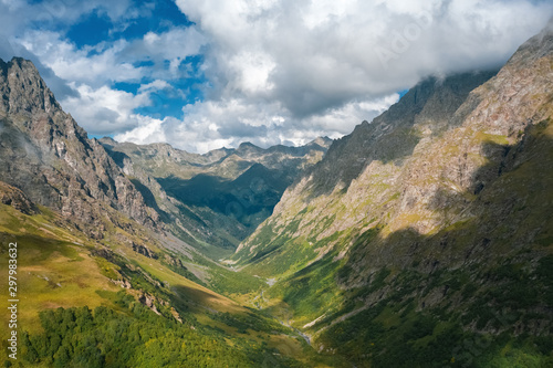 Beautiful river valley with mountain ranges on the sides with steep stony slopes overgrowth by alpine meadows and green forest; early autumn day with overcast sky; popular touristic route, Caucasus