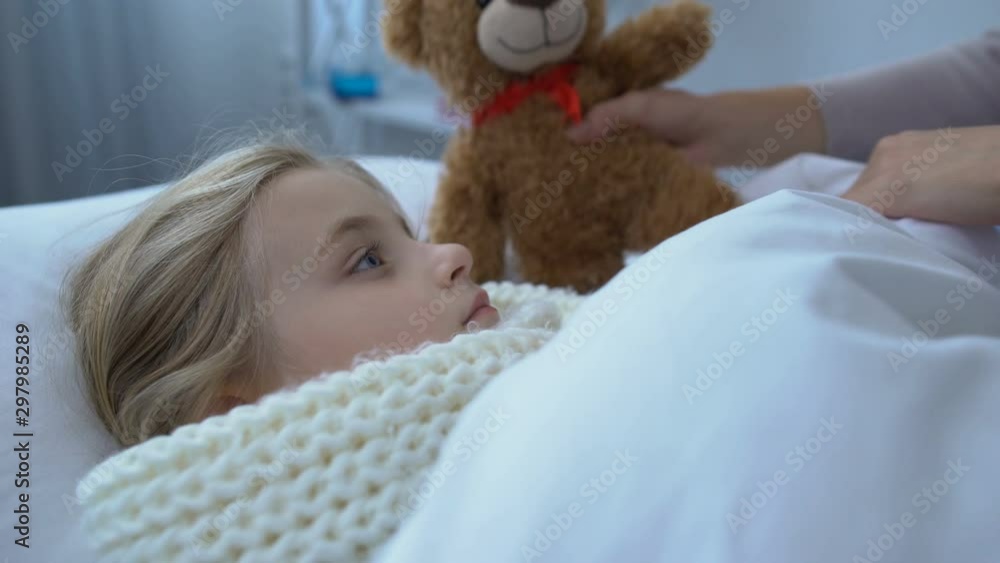 Sick female kid in scarf lying in bed, looking at mom giving teddy bear epidemic Sick female kid in scarf lying in bed, looking at mom giving teddy bear epidemic