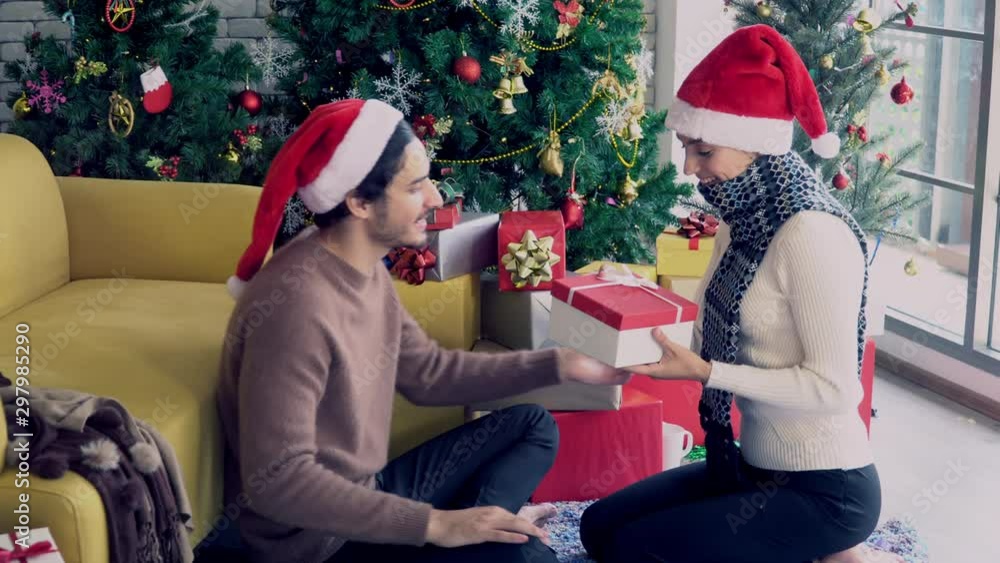 4K Wide shot of beautiful woman girlfriend wearing santa hat give christmas gift to her boyfriend with Christmas tree background. Happy family celebration festival, love and romantic moment concept. 4K Wide shot of beautiful woman girlfriend wearing santa hat give christmas gift to her boyfriend with Christmas tree background. Happy family celebration festival, love and romantic moment concept.
