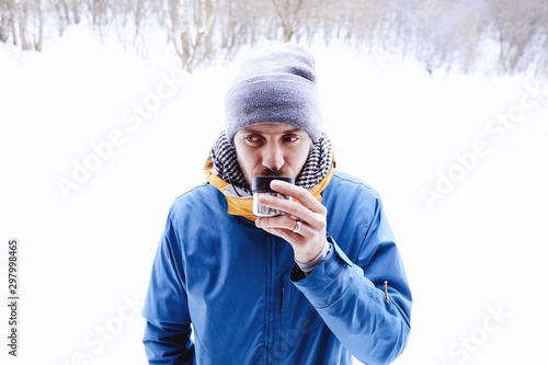 A young bearded man in a hat and a blue jacket drinks tea coffee outdoors on a winter day. White background