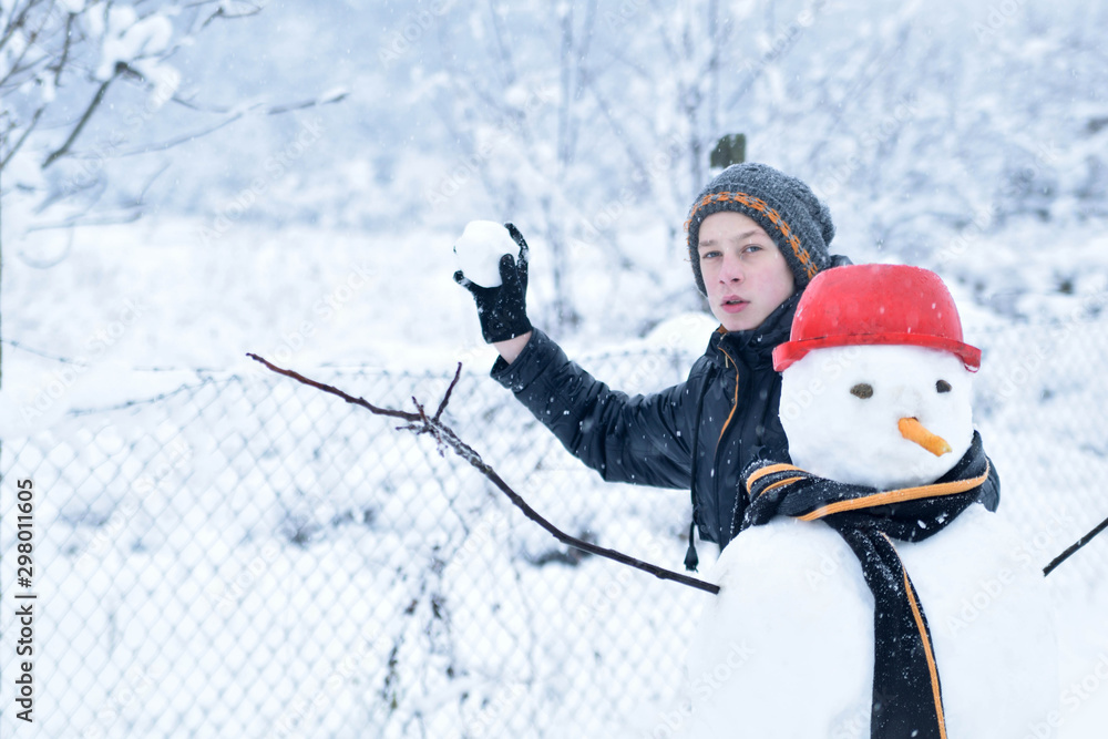 Fototapeta premium Winter teenager in a jacket and hat sculpts a snowman on a winter background with falling snow in frozen day concept
