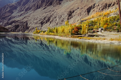 Small village in Gupis Ghizer valley with a view of reflection in still water of blue calmness Khalti lake against colorful trees in autumn season and Hindu Kush mountain. Gilgit Baltistan, Pakistan.