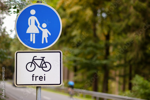 A round blue and white sign on a forest road indicates a path for pedestrians. Cycling is on the road. A white sign says: bicycles free. Translation of the sign: free