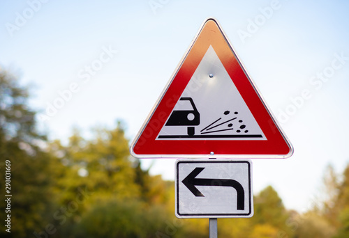 A triangular red white warning sign. The road is under construction and there are small stones and granules on the road. In the background, green trees out of focus with beautiful bokeh.