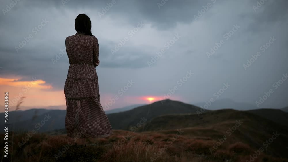 Young mysterious girl in a long dress stands on top of a mountain and enjoys the view. Dramatic mountain landscape with red sunset before rain and thunderstorm Young mysterious girl in a long dress stands on top of a mountain and enjoys the view. Dramatic mountain landscape with red sunset before rain and thunderstorm