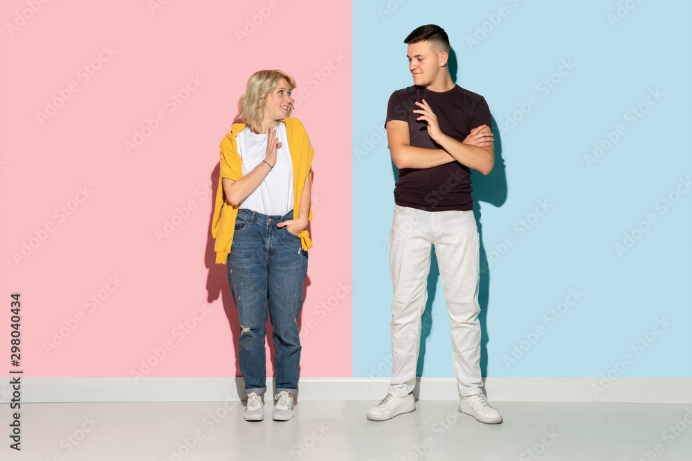 Young emotional man and woman in bright casual clothes posing on pink and blue background. Concept of human emotions, facial expession, relations, ad. Beautiful caucasian couple greeting and smiling.