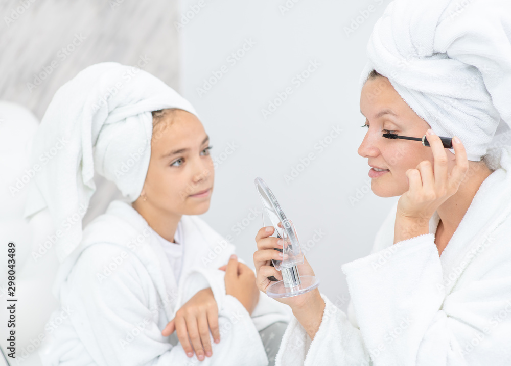 Little girl learns from mom to apply makeup at home. Mother and child girl are in bathrobes and with towels on their heads Little girl learns from mom to apply makeup at home. Mother and child girl are in bathrobes and with towels on their heads