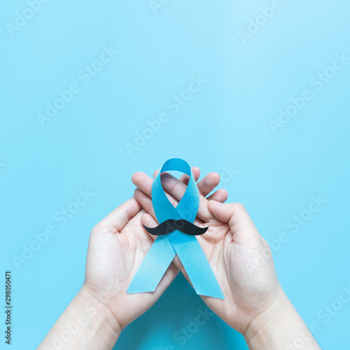 Men's health and Prostate cancer awareness campaign in November. Man hands holding light blue ribbon awareness w/ mustache on blue background. Symbol for support men who living w/ cancer. Copy space.