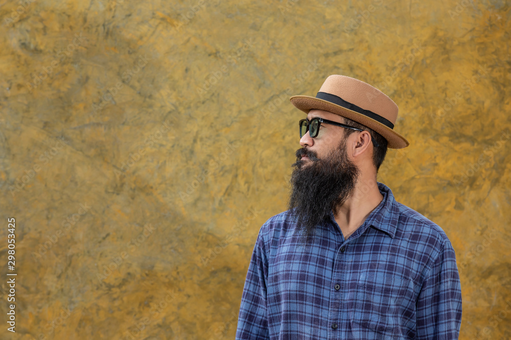 Movember Concept. Handsome young man with a long beard wearing a long-sleeved blue shirt, wearing a hat and glasses standing on a yellow cement background.
