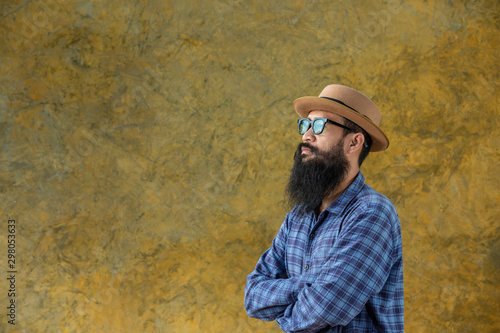 Movember Concept. Handsome young man with a long beard wearing a long-sleeved blue shirt, wearing a hat and glasses standing on a yellow cement background. © artit