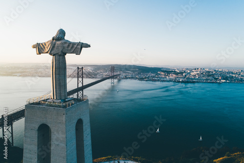 Aeria view monument Sanctuary of Christ the King. Drone flyby past near giant sculpture overlooking city of Lisbon Almada and famous 25th april bridge over river Tagus at sunset photo
