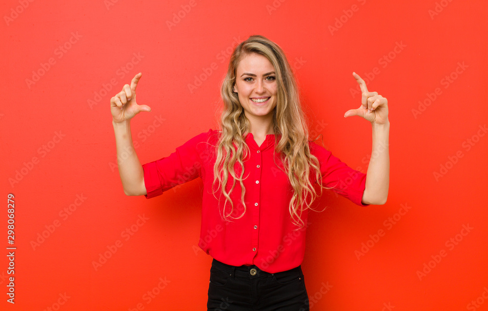 young blonde woman framing or outlining own smile with both hands, looking positive and happy, wellness concept against red wall - obrazy, fototapety, plakaty 