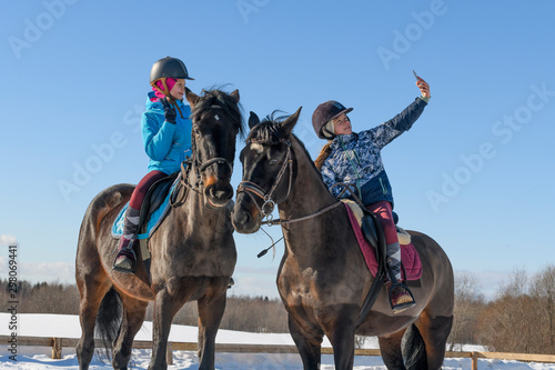 Two caucasian female teenager, 13 years old, are astride their horses and taking a selfie with one smartphone on the training arena in winter.