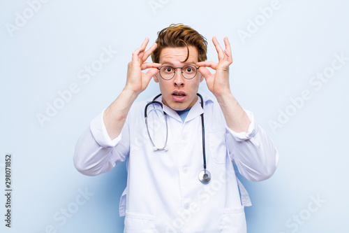 young red head doctor feeling shocked, amazed and surprised, holding glasses with astonished, disbelieving look against blue wall photo