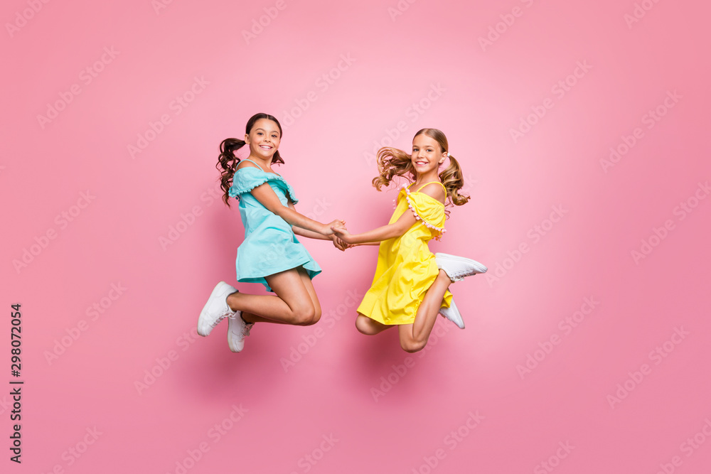 Full body profile photo of two small sisters jumping high together last studying day holding hands best friends wear blue yellow dresses isolated pink color background Full body profile photo of two small sisters jumping high together last studying day holding hands best friends wear blue yellow dresses isolated pink color background
