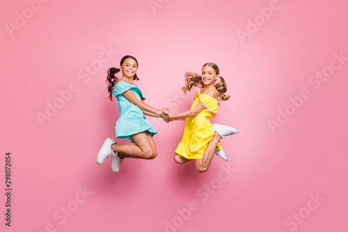 Full body profile photo of two small sisters jumping high together last studying day holding hands best friends wear blue yellow dresses isolated pink color background